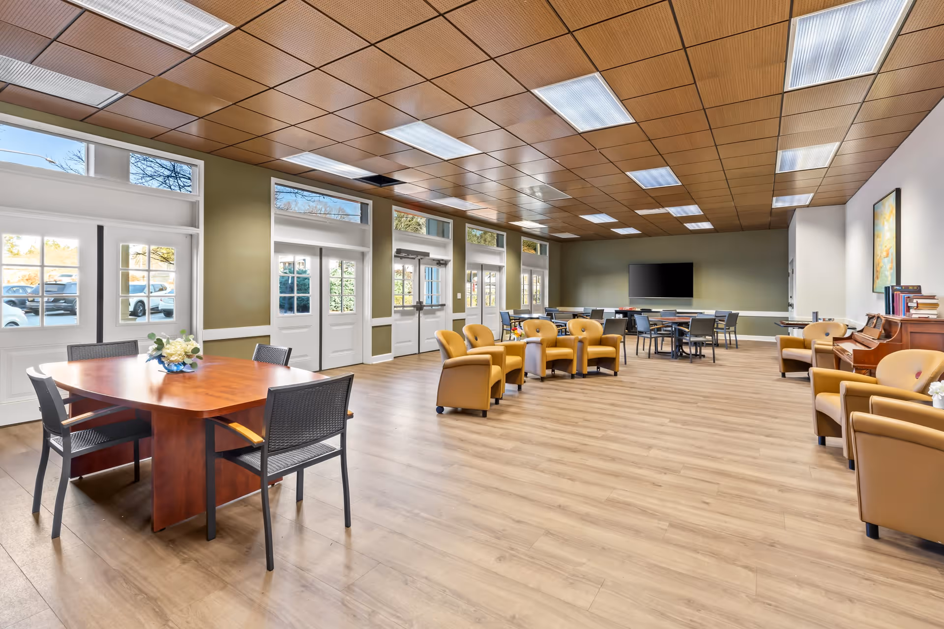 A spacious senior living facility common area with wooden flooring and a wooden paneled ceiling. The room features multiple seating arrangements including a round wooden table with four chairs, several yellow armchairs arranged in a semi-circle, and additional tables and chairs towards the back near a large flat-screen TV mounted on the wall. There is a piano on the right side of the room and large windows and doors along the left wall allowing natural light to enter.