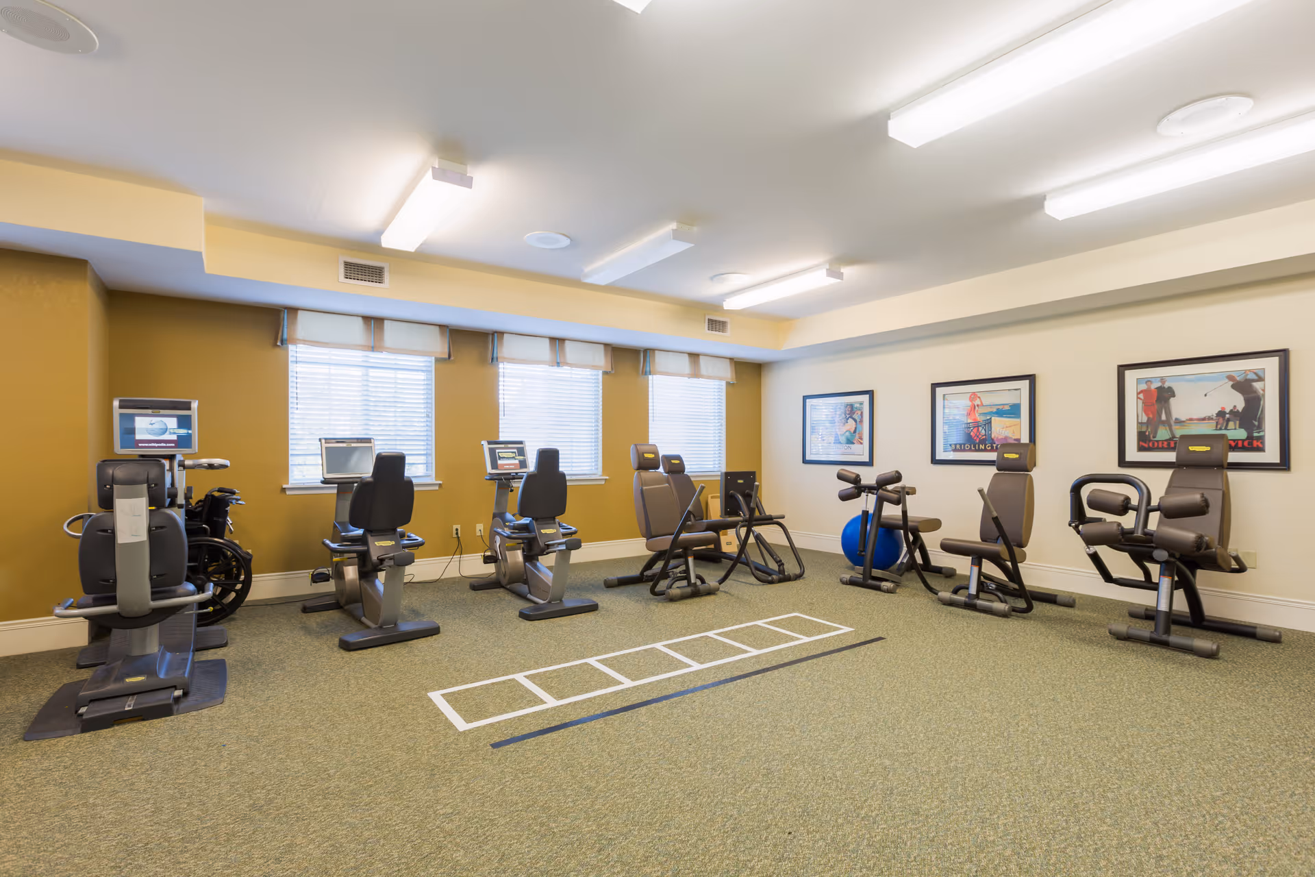 A well-lit exercise room with various fitness equipment including stationary bikes, leg press machines, and other workout chairs. The room has three windows with blinds, framed fitness-themed posters on the wall, and a carpeted floor with a white ladder agility training marking.