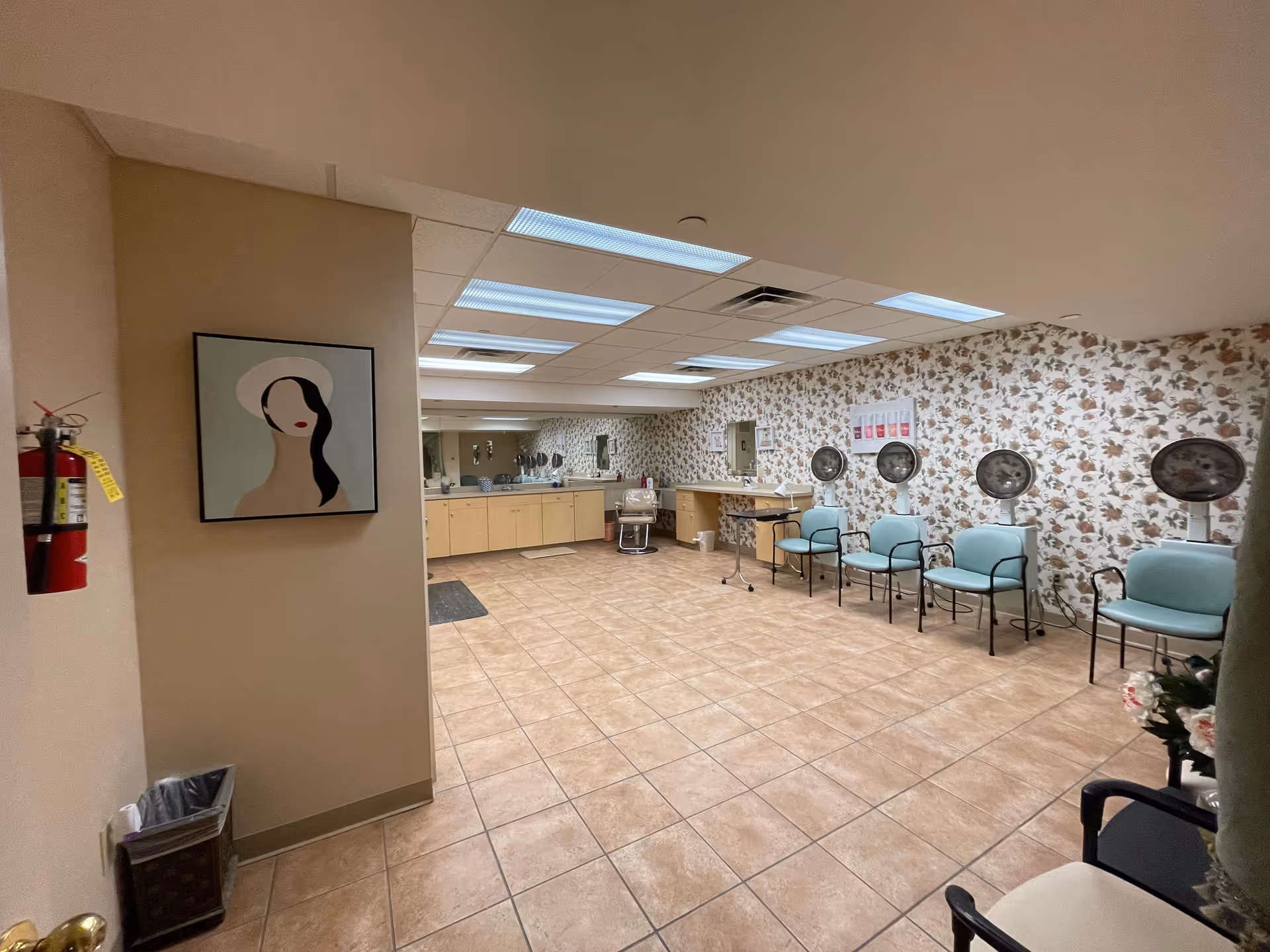 Interior view of a salon area with floral wallpaper, four blue salon chairs with hair dryers above them, a counter with cabinets and a mirror along the back wall, a fire extinguisher on the left wall, and a framed abstract portrait painting.