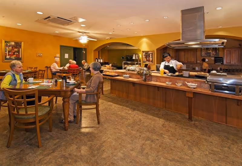 Residents sit at tables in a dining area while a staff member prepares food behind a long serving counter.