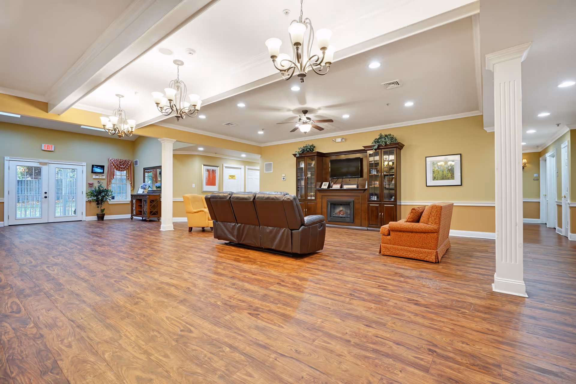 A spacious living room area in a senior living facility with wood flooring, a brown leather sofa, an orange armchair, and a yellow armchair. The room features a large wooden entertainment center with a TV and a fireplace, ceiling fans, chandeliers, and light yellow walls with white trim. Double glass doors lead outside, and there are decorative plants and framed artwork on the walls.
