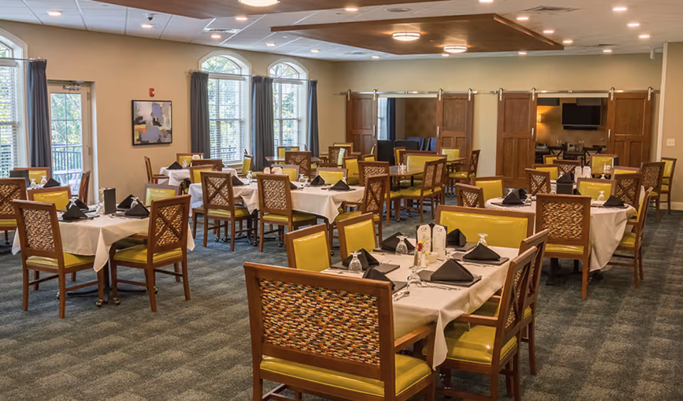 A spacious dining room with multiple tables covered in white tablecloths, each set with black folded napkins, glasses, and menus. The chairs have wooden frames with patterned backs and yellow cushioned seats. Large windows with gray curtains allow natural light to fill the room. The room has a carpeted floor and a ceiling with recessed lighting.