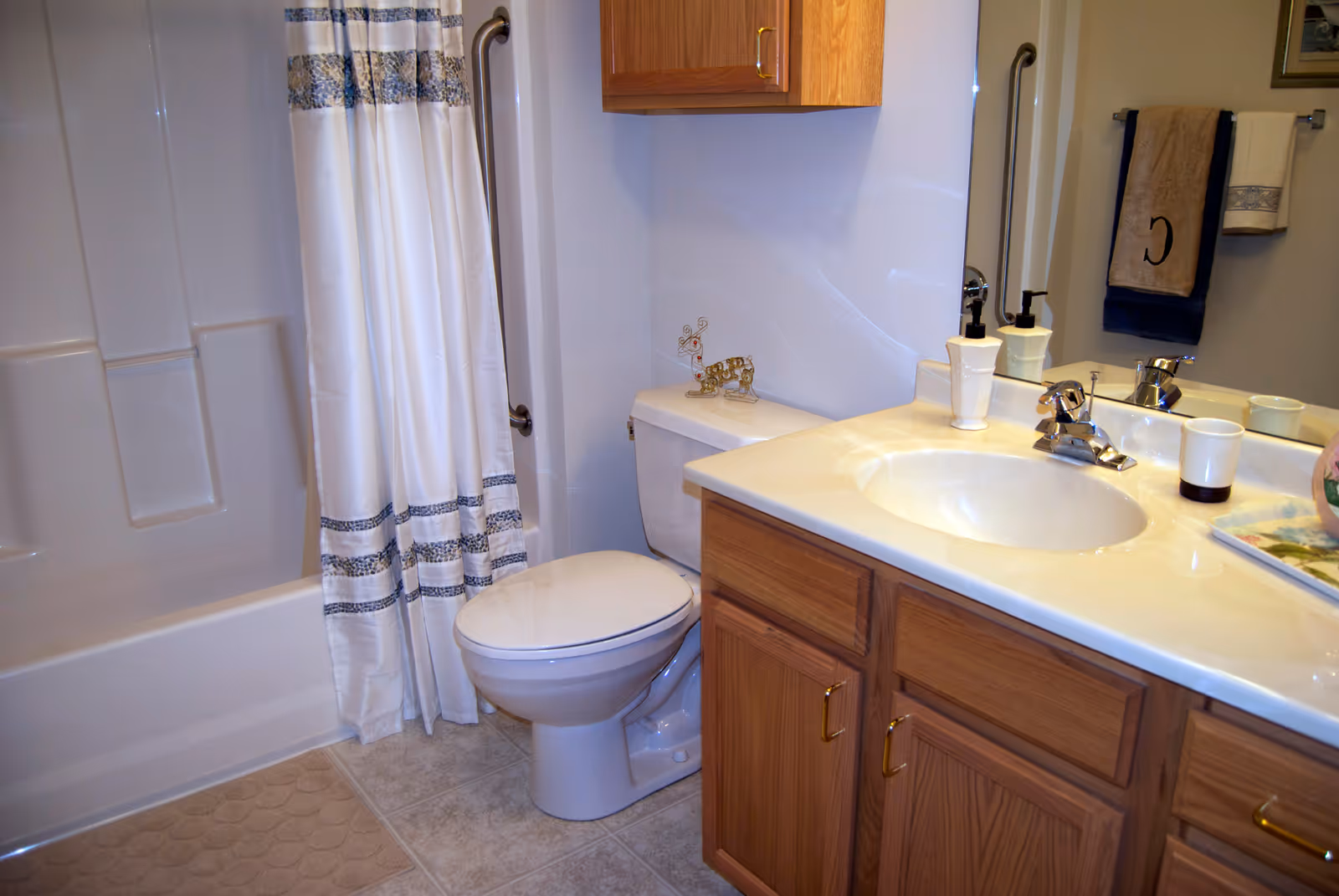 A clean bathroom featuring a white toilet, a bathtub with a white shower curtain decorated with blue patterns, a beige bath mat, a wooden vanity with a white countertop and sink, a mirror above the sink, and a towel rack with beige and blue towels.