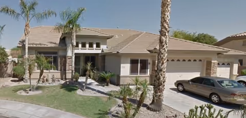 Exterior view of a single-story residential building with a tiled roof, a three-car garage, palm trees, and a car parked in the driveway under a clear blue sky.