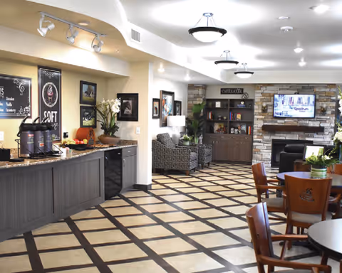 Bright common area with a coffee station, round dining tables, lounge seating, and a TV over a fireplace.