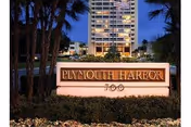Illuminated Plymouth Harbor sign in foreground with a tall building and palm trees behind.