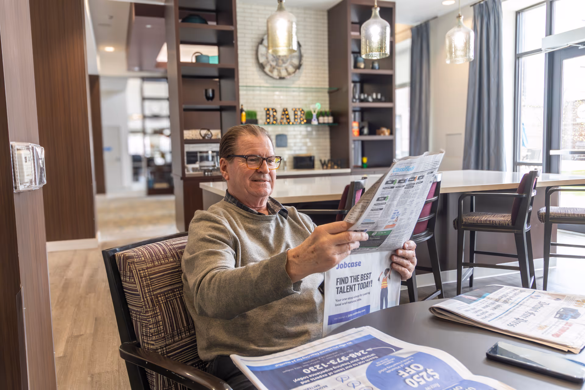 An older man sits at a table in a communal dining area reading a newspaper with a kitchen/bar and pendant lights in the background.