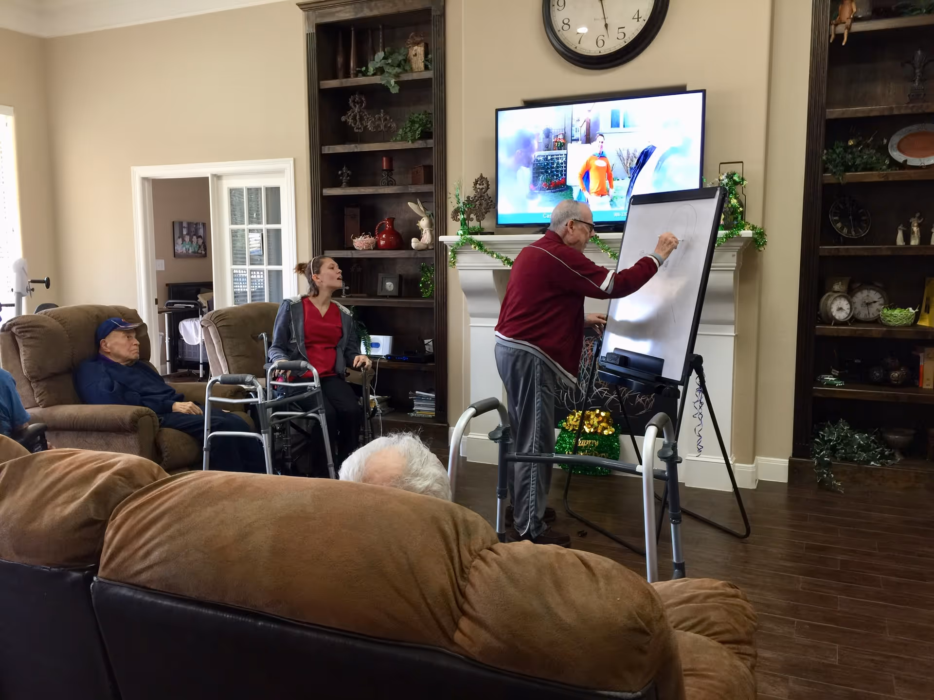 Several seniors sit in a living room while one man draws on an easel in front of a TV and fireplace.
