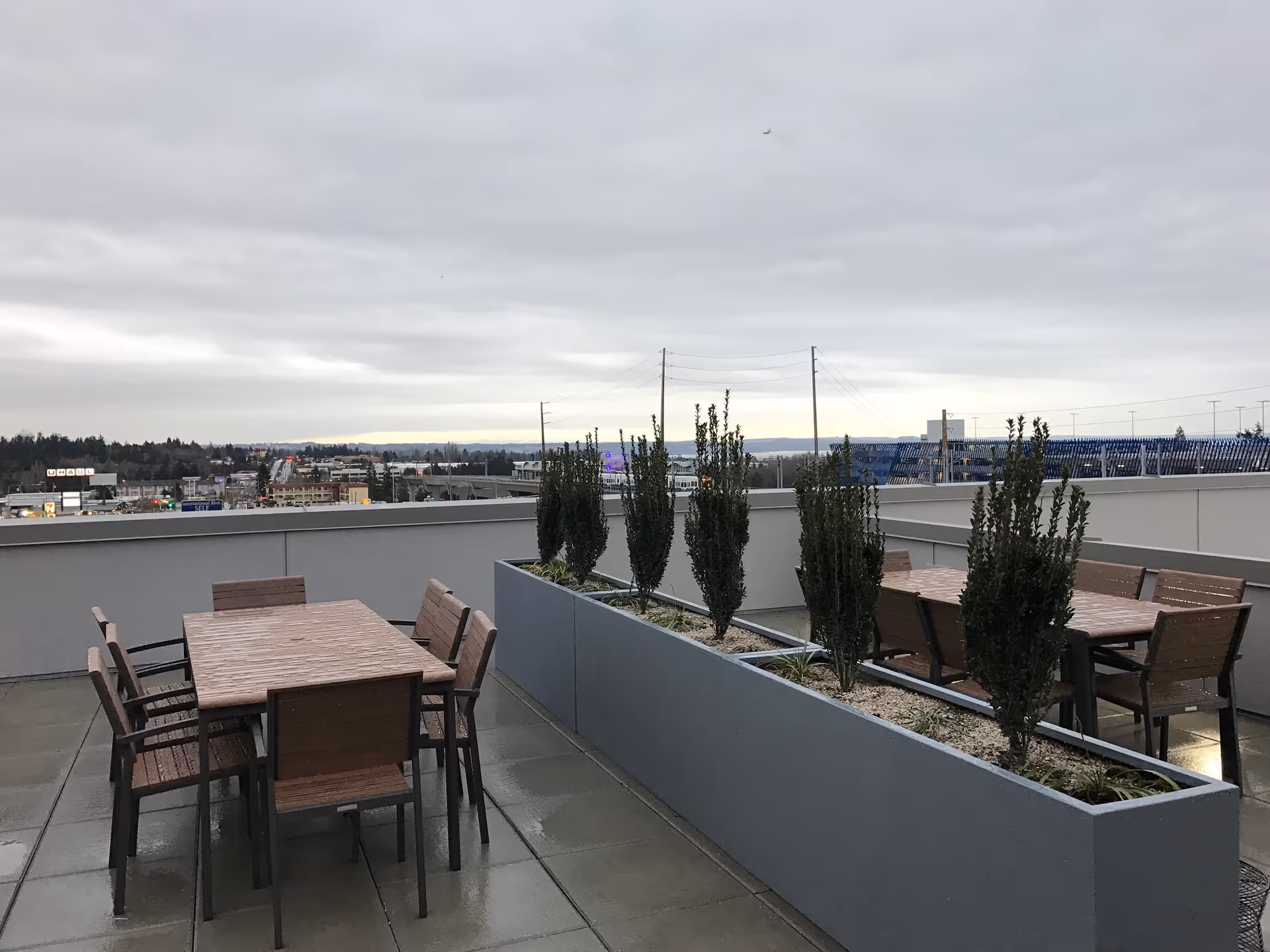 Outdoor rooftop patio area with two wooden tables and several chairs around them. A long rectangular planter with tall green plants separates the tables. The sky is overcast and a cityscape with buildings and trees is visible in the background.