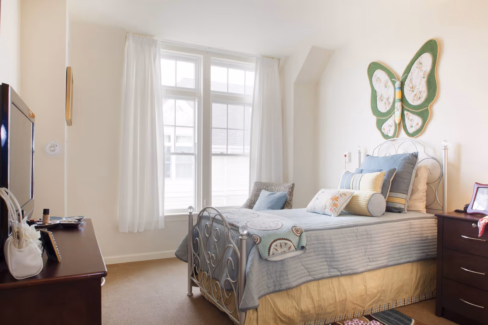 A bright bedroom with a single bed featuring a metal frame, light blue bedding, and multiple pillows. There is a large butterfly wall decoration above the bed. The room has a window with white curtains letting in natural light. A dark wooden dresser with a TV and personal items is on one side of the room.