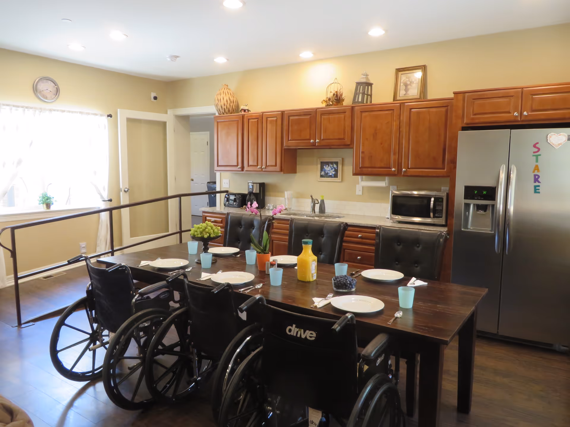 A kitchen and dining area with a wooden table set for six people. There are three wheelchairs on one side of the table and three cushioned chairs on the other side. The table has plates, cups, cutlery, a bottle of orange juice, grapes, and a potted orchid. The kitchen has wooden cabinets, a stainless steel refrigerator with colorful magnets, a microwave, and a coffee maker. A window with sheer curtains lets in natural light.