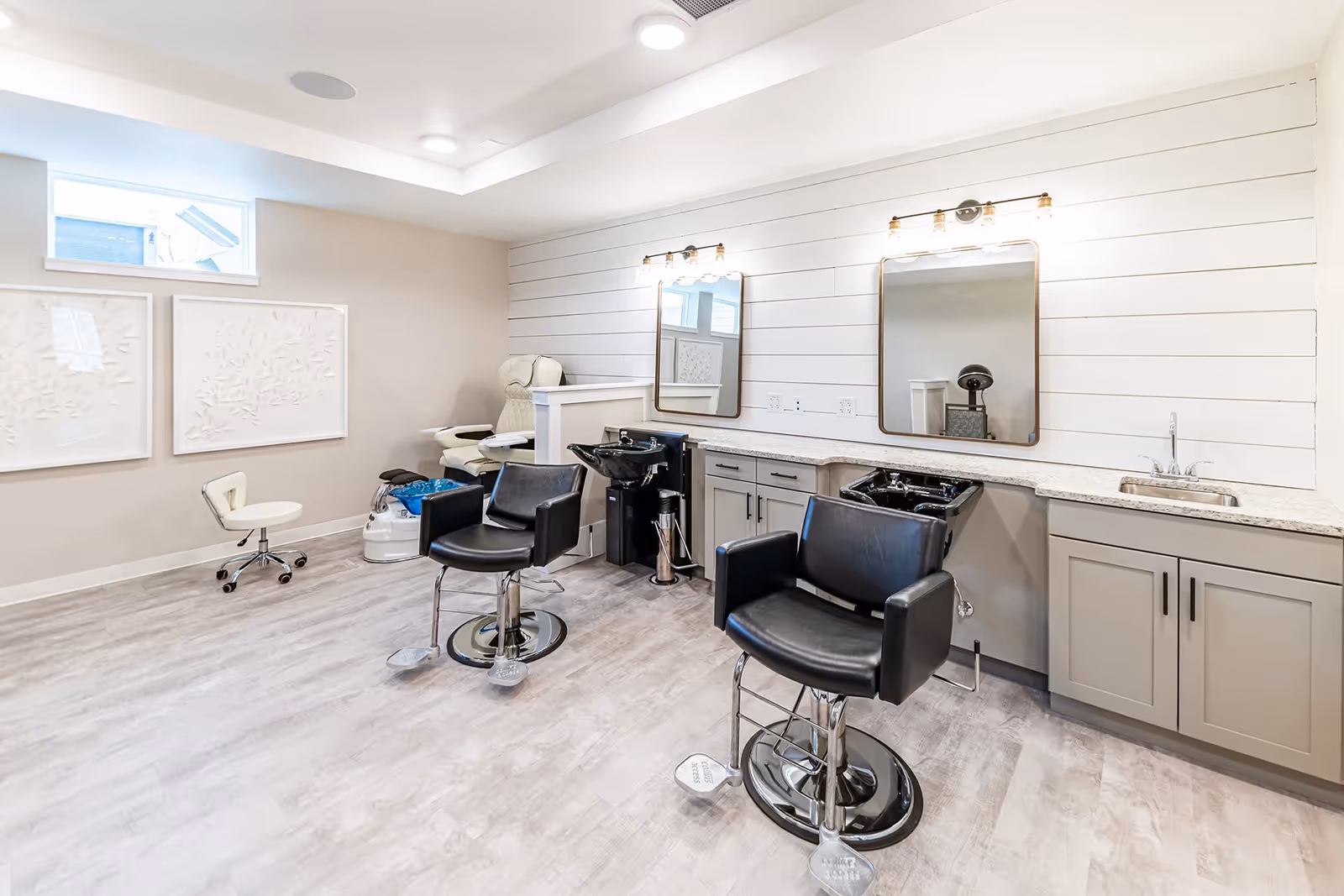 A bright and clean salon area in a senior living facility featuring two black salon chairs in front of two mirrors mounted on a white shiplap wall. Below the mirrors are two black hair washing sinks integrated into a countertop with cabinets. To the left, there is a white pedicure chair and a small rolling stool. The room has light-colored wood flooring and soft beige walls with two decorative white framed artworks. A small window near the ceiling allows natural light in.