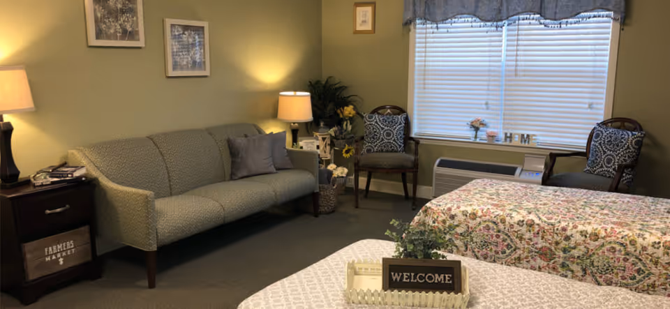 A cozy living room area with a green patterned sofa, two wooden chairs with blue patterned cushions, a side table with a lamp, and decorative plants. There are two beds with floral bedspreads in the foreground, a window with blinds and a valance, and a small sign that says 'WELCOME' on a tray on one of the beds.