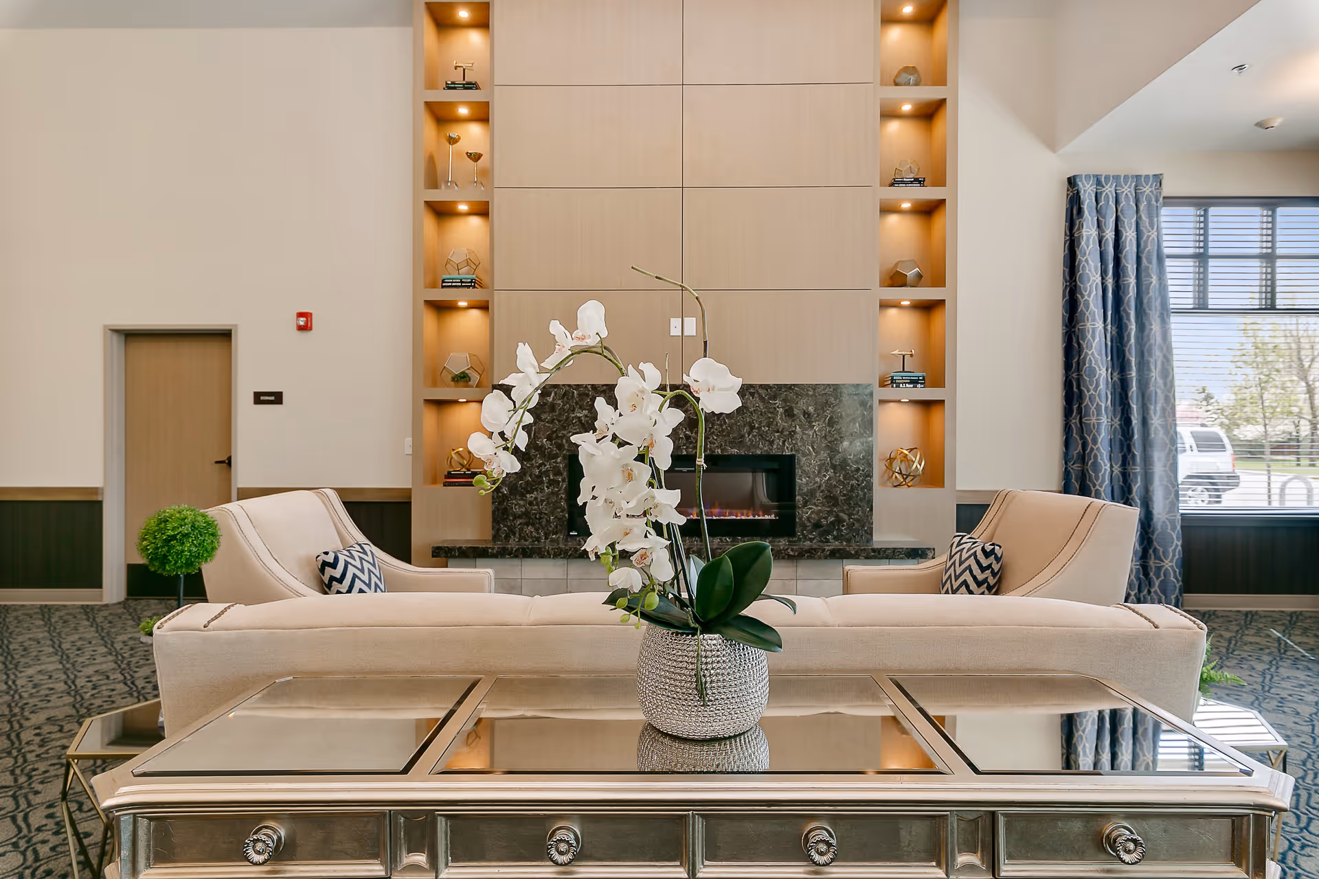 A modern senior living facility lounge area featuring a beige sofa with patterned pillows facing a decorative fireplace with built-in shelves illuminated by recessed lighting. A glass-top coffee table with a silver vase holding white orchids is in the foreground. Large windows with blue patterned curtains allow natural light into the room.