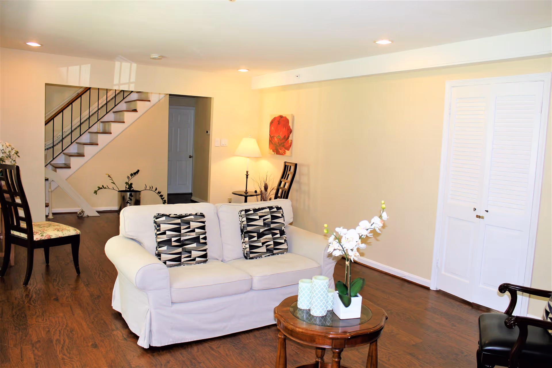 A bright living room with a white loveseat featuring two black and white patterned pillows, a small round wooden coffee table with decorative items including a white orchid plant and candles, wooden flooring, a staircase with a black railing in the background, a floor lamp next to a chair, and a closed white double door on the right wall.