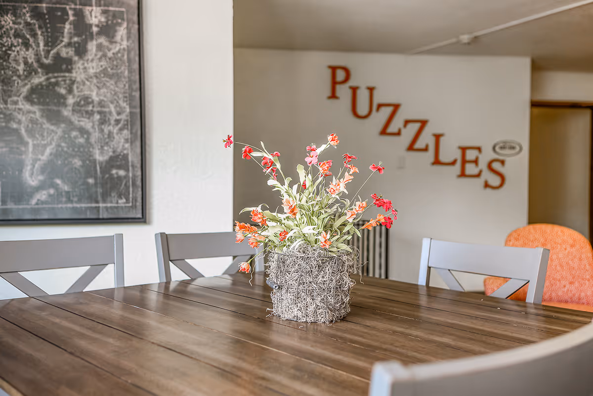 A wooden table with a decorative flower arrangement in a woven basket sits in a room with gray chairs around it. On the wall in the background, the word 'PUZZLES' is displayed in large red letters. A framed map hangs on the left wall, and an orange chair is partially visible on the right.