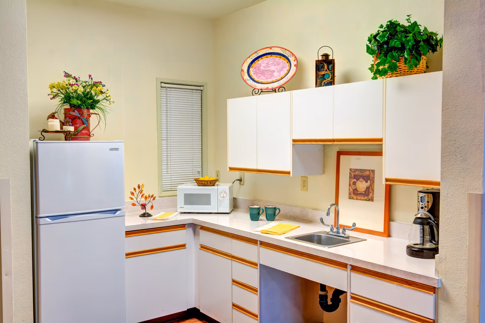 A small kitchen area with white cabinets and wooden handles, a white refrigerator, a microwave, a coffee maker, and a stainless steel sink. The countertop has two green mugs, a yellow napkin, and decorative items including a framed picture, a colorful plate, and plants.