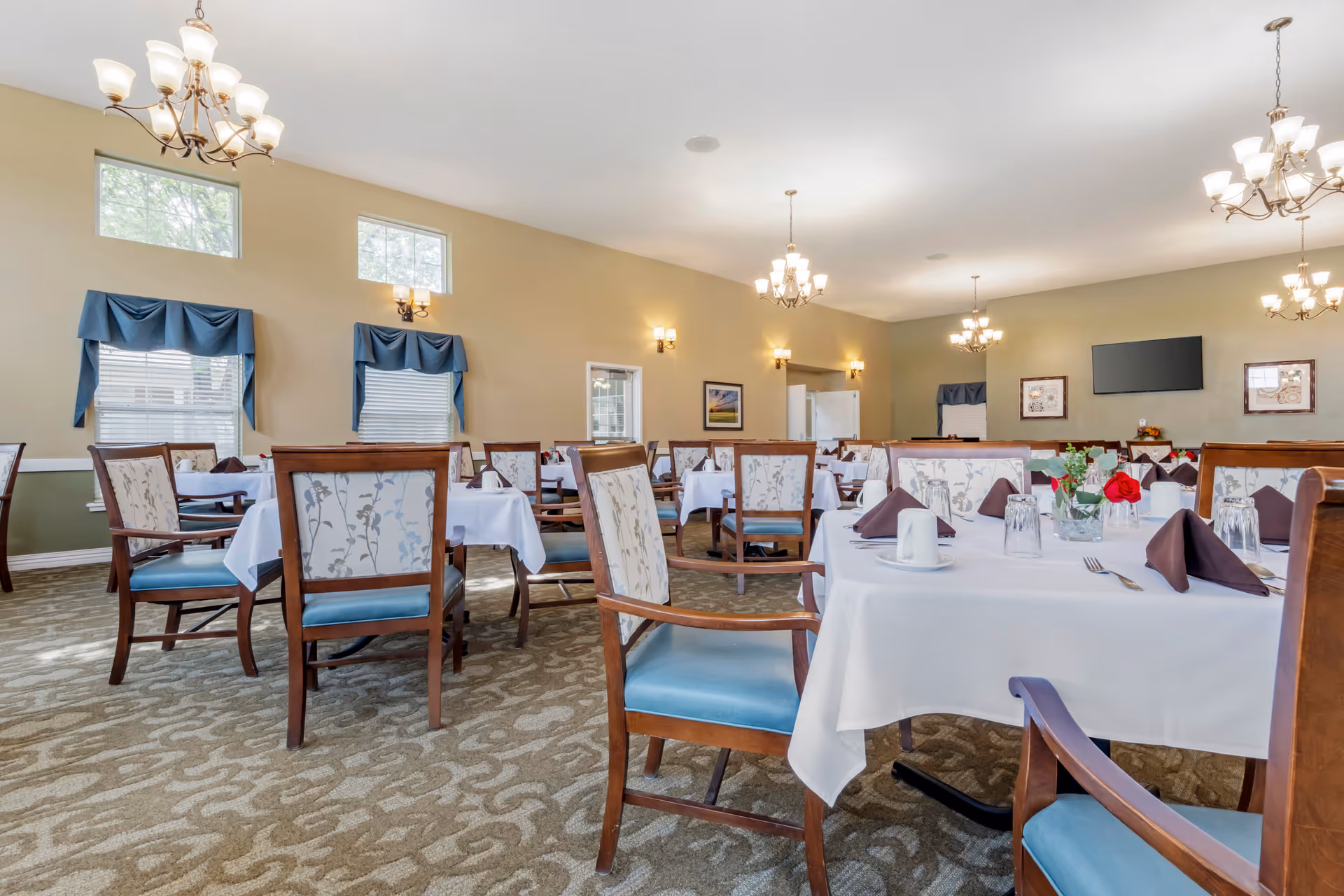 A spacious dining room with multiple tables covered in white tablecloths, each set with brown napkins, cups, glasses, and silverware. The chairs have wooden frames with blue cushioned seats and floral-patterned backs. The room is well-lit with chandeliers and wall sconces, and there are windows with blue valances letting in natural light. A flat-screen TV and framed pictures are mounted on the far wall.