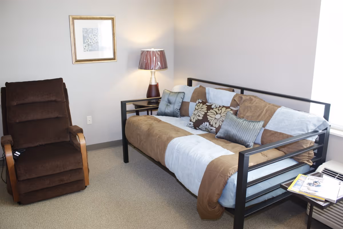 A cozy room with a brown cushioned recliner chair, a daybed with a patchwork brown and white comforter and decorative pillows, a small side table with a lamp, a framed floral picture on the wall, and a small table holding magazines near a window.