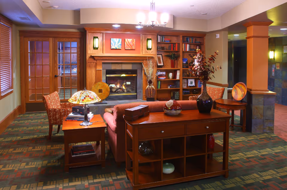 Cozy living room area with a lit fireplace surrounded by wooden shelves filled with books and decorative items. There is a red sofa facing away from the fireplace, a wooden table behind it with vases and decorative objects, and a side table with a stained glass lamp and books. Two patterned armchairs are positioned near the fireplace and the room has warm lighting and carpeted floors with a geometric pattern.