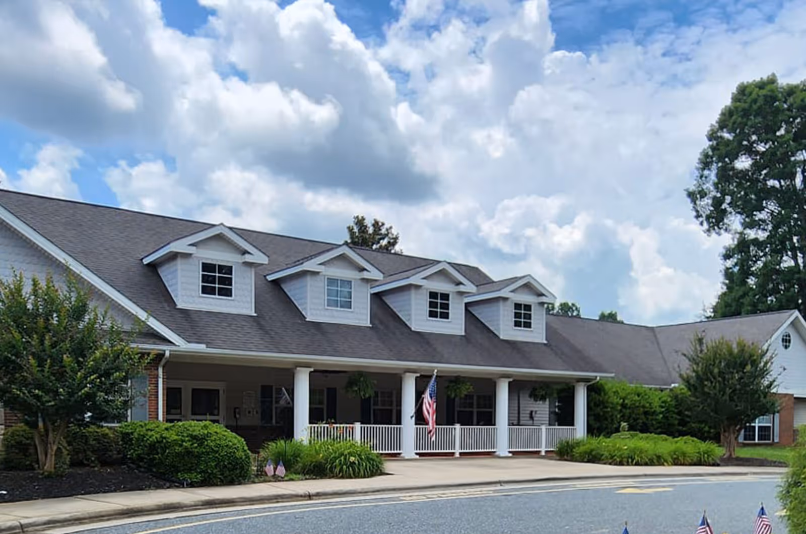 Front exterior view of Ramseur Rehabilitation and Healthcare Center, a single-story building with a gray roof, white siding, and three dormer windows. The entrance features a covered porch with white columns and an American flag displayed. There are bushes and small trees around the building, and a partly cloudy sky overhead.