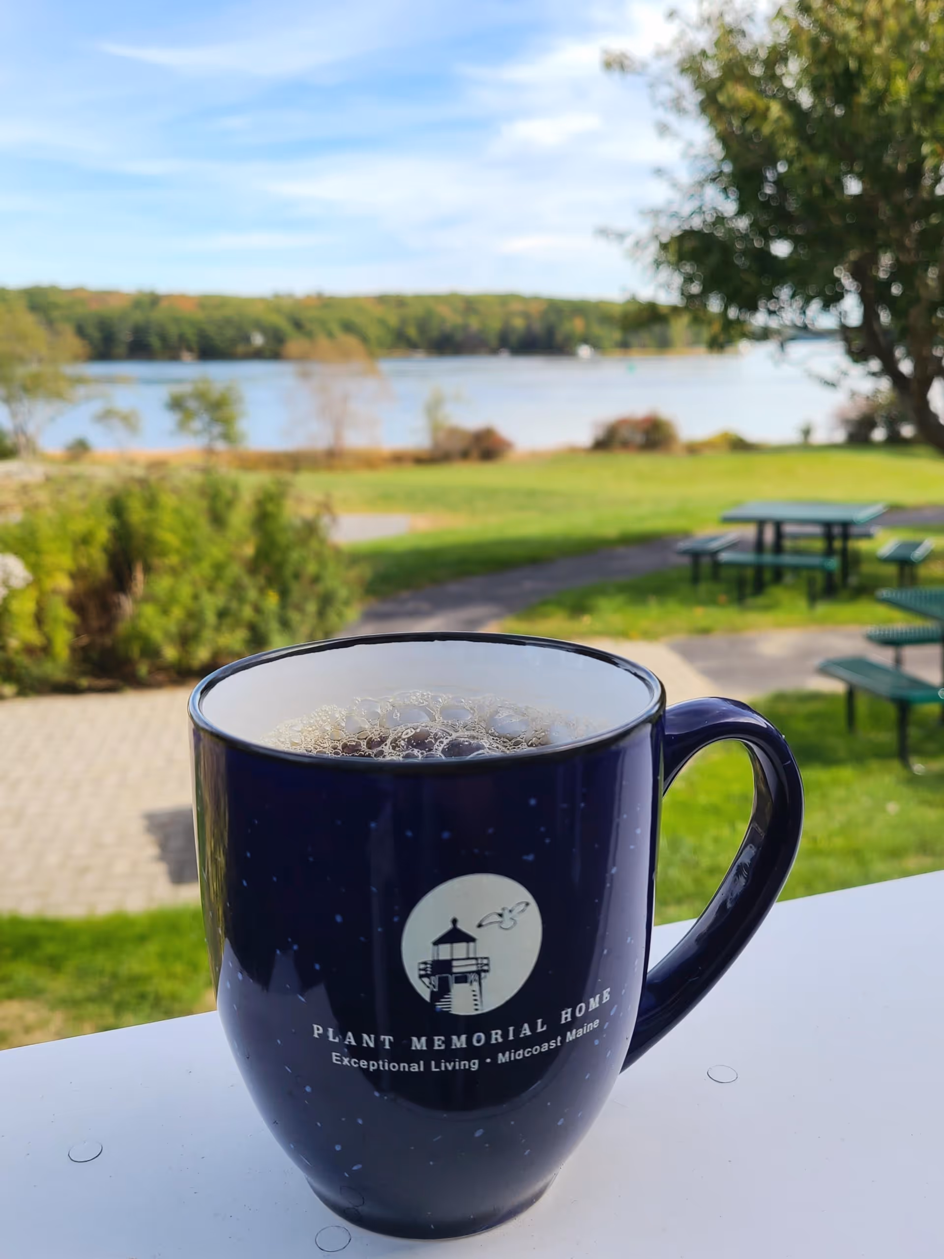 A dark blue coffee mug with the logo and text 'Plant Memorial Home Exceptional Living Midcoast Maine' sits on a white railing. In the background, there is a green lawn with picnic tables, trees, and a body of water under a partly cloudy sky.