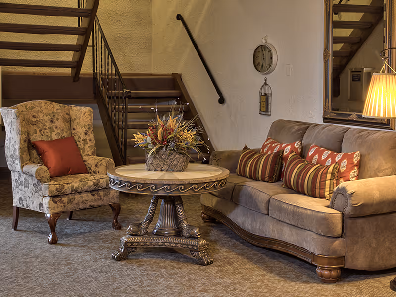 Cozy seating area with a tufted sofa, patterned armchair, ornate round coffee table with a floral centerpiece, and a staircase in the background.