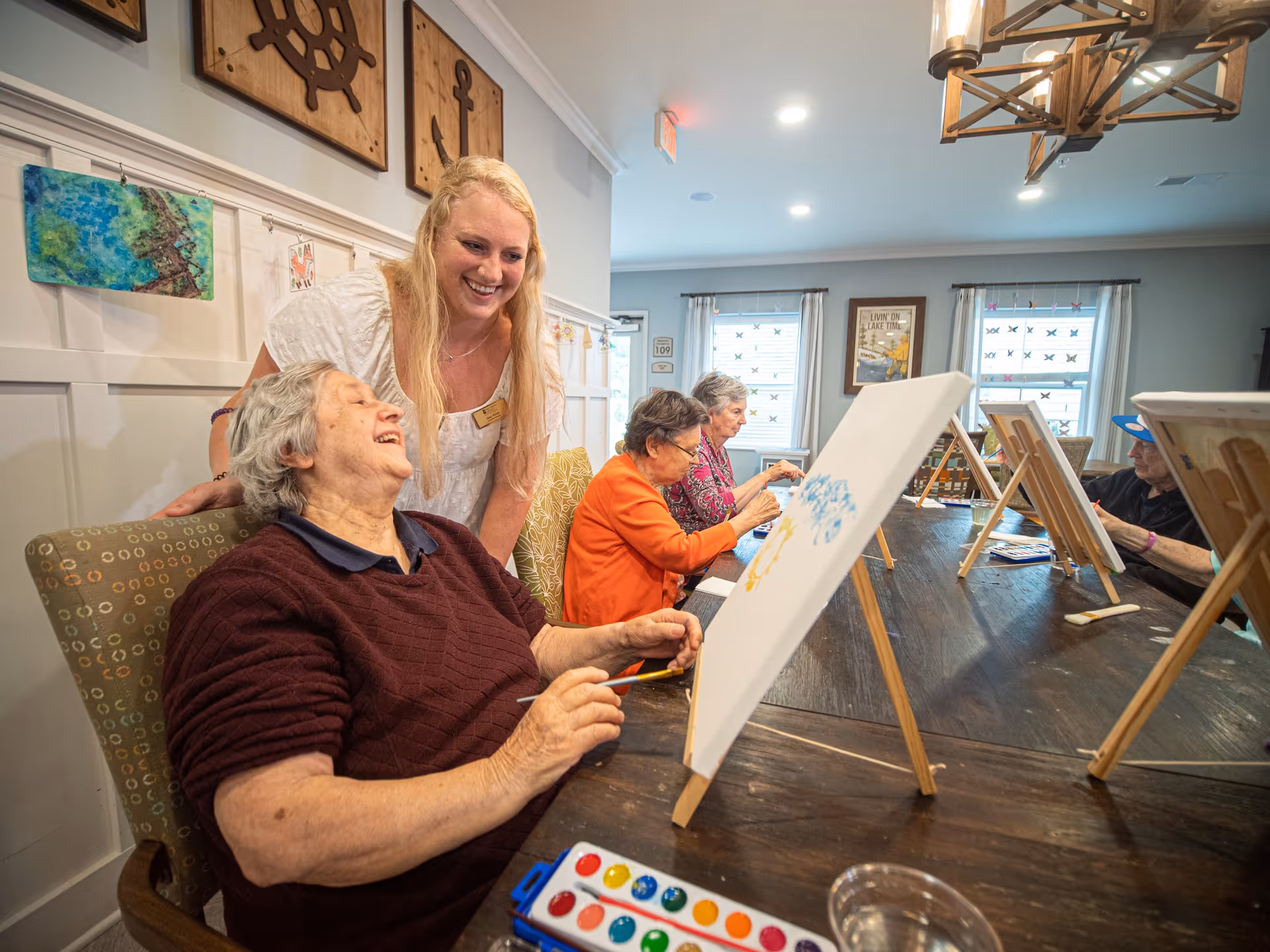 A group of elderly women seated around a table engaged in a painting activity with canvases on easels. A smiling caregiver or instructor leans over one woman who is laughing. The room is well-lit with windows and decorated with nautical-themed wall art.