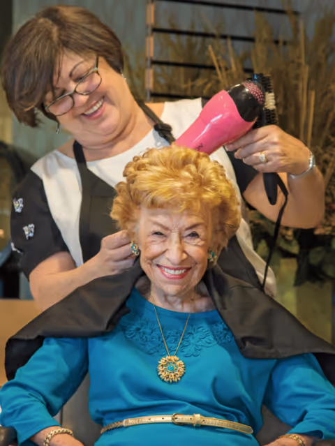 An elderly woman with styled blonde hair and a bright smile is seated while a hairstylist uses a pink hairdryer and brush to style her hair. The hairstylist is smiling and wearing glasses and a black apron. The setting appears to be a salon or hair care area within a senior living facility.