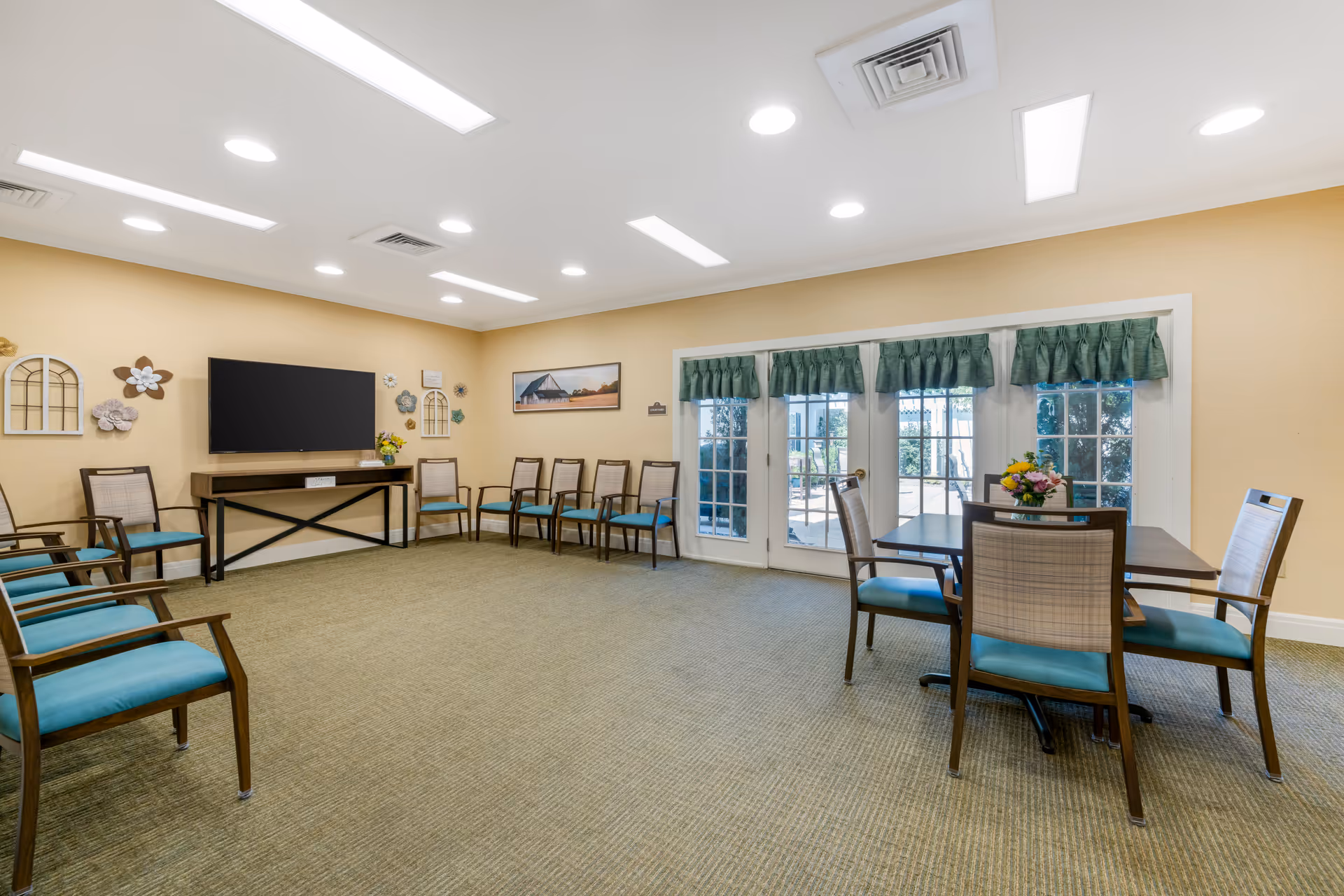 A spacious common room with beige walls and carpeted floor. The room features a row of wooden chairs with teal cushions lined up against the wall, a flat-screen TV mounted on the wall above a console table, and a small table with four chairs near glass doors with green valance curtains that lead outside. Decorative wall art and flowers add a welcoming touch.