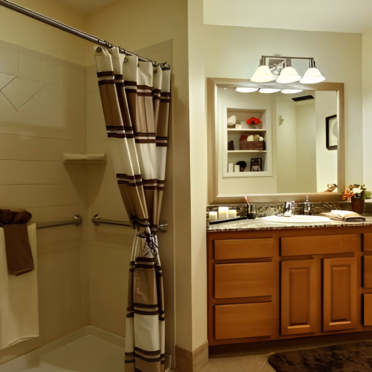 A bathroom with a shower area featuring a striped shower curtain and grab bars, a towel hanging on a rack, and a wooden vanity with a granite countertop, sink, and a large mirror above it. The mirror reflects shelves with decorative items and a light fixture with three lamps.