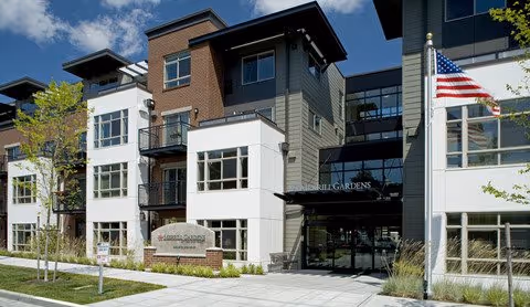 Exterior view of a modern multi-story senior living facility building with a combination of brick and light-colored siding. The entrance is visible with a sign that reads Merrill Gardens. There are small trees and landscaping along the sidewalk, and an American flag is displayed on the right side of the building.