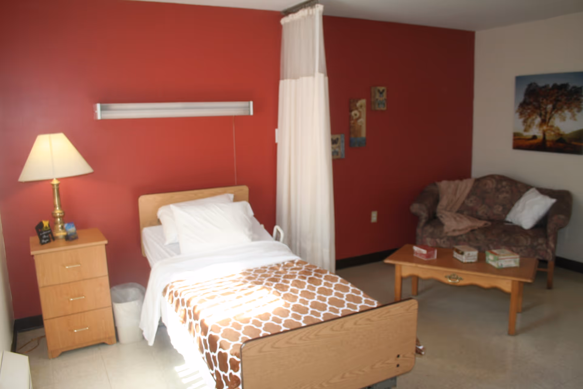 A senior care facility room with a single bed covered with white sheets and a brown patterned blanket. Next to the bed is a wooden nightstand with a lamp and some small items. A white privacy curtain hangs beside the bed. In the background, there is a floral-patterned loveseat with a pillow and blanket, a wooden coffee table with tissue boxes, and a wall decorated with small paintings and a large picture of a tree at sunset. The walls are painted red and white.
