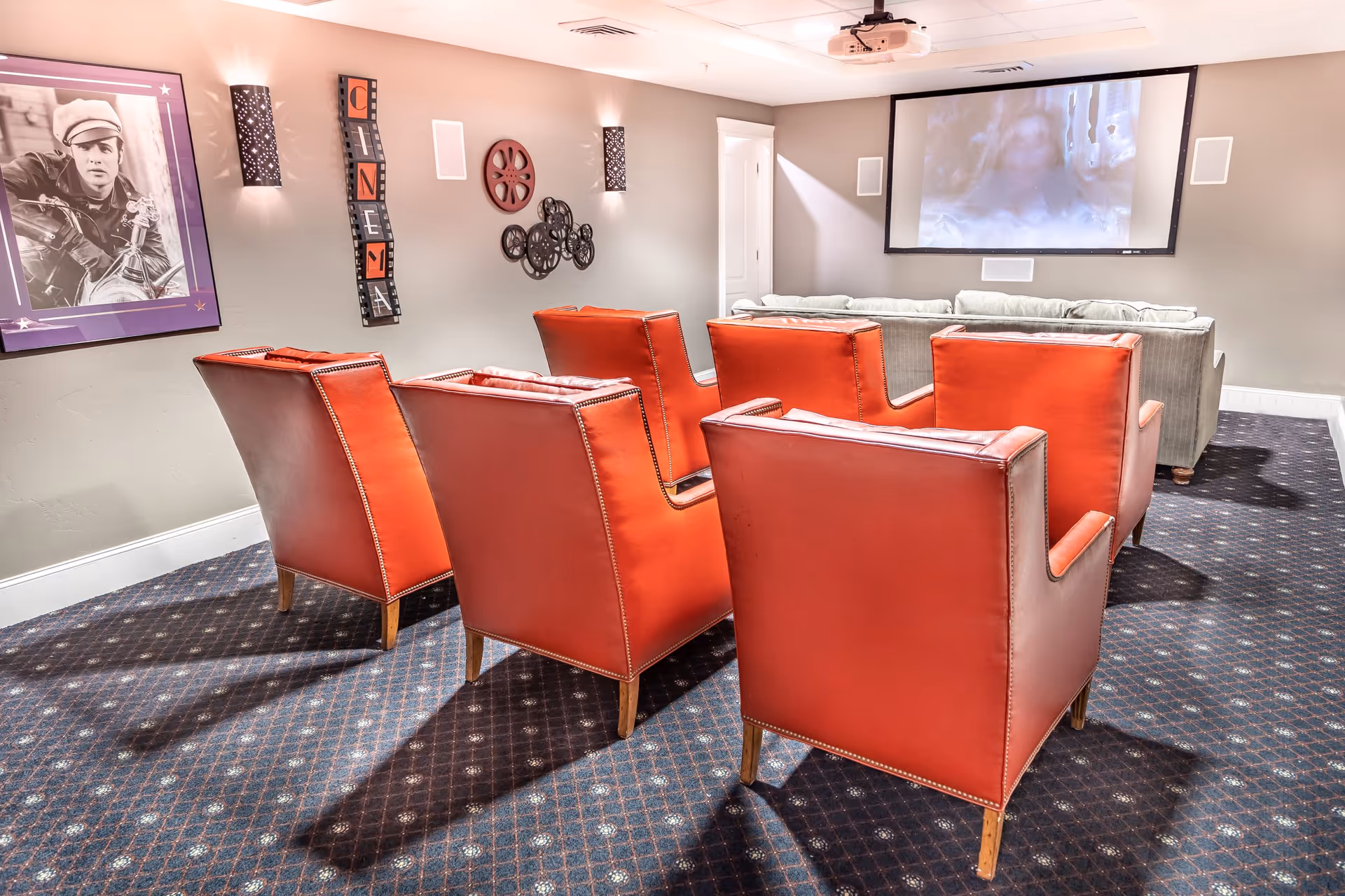 A cozy home theater room with six red leather armchairs arranged in two rows facing a large screen on the wall. The room has patterned carpet flooring, gray walls decorated with movie-themed wall art including a framed black and white photo, film reel decorations, and a vertical sign spelling 'CINEMA'. There is a gray couch behind the armchairs and ceiling-mounted projector above.