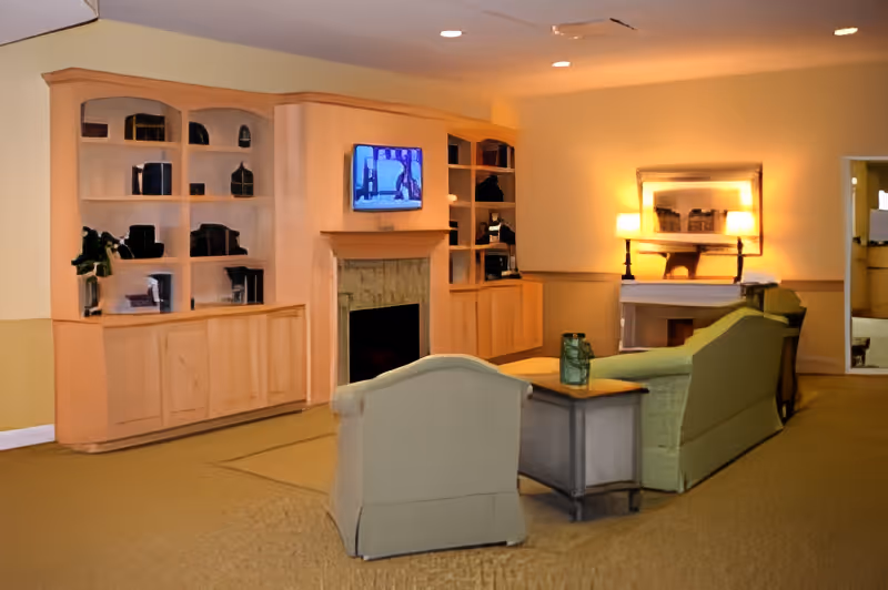Common living area with upholstered chairs and a sofa arranged around a fireplace and wall-mounted TV, with built-in wooden shelving and warm lighting.