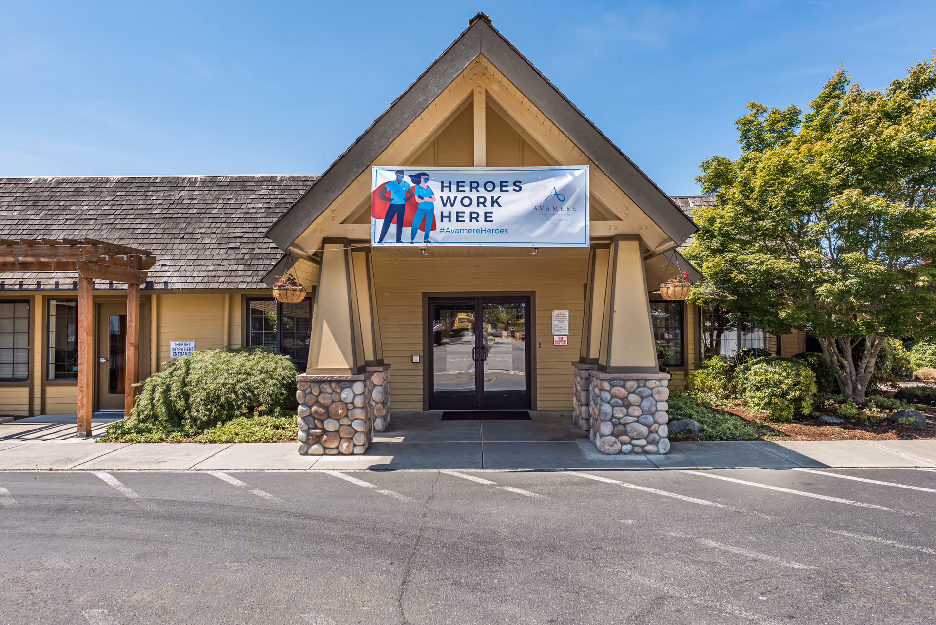 Front entrance of Avamere Olympic Rehabilitation of Sequim building with a peaked roof supported by stone pillars. A banner above the entrance reads 'HEROES WORK HERE #AvamereHeroes'. There are bushes and trees on either side of the entrance and a clear blue sky above.