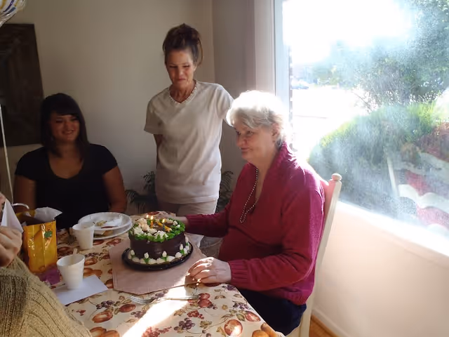 Three women gathered around a table with a chocolate cake decorated with white flowers. One woman in a red sweater is seated, while another woman in a white shirt stands behind her, and a third woman in a black shirt is seated to the left. The table is covered with a patterned tablecloth and has cups and plates on it. Sunlight is coming through a window on the right side.