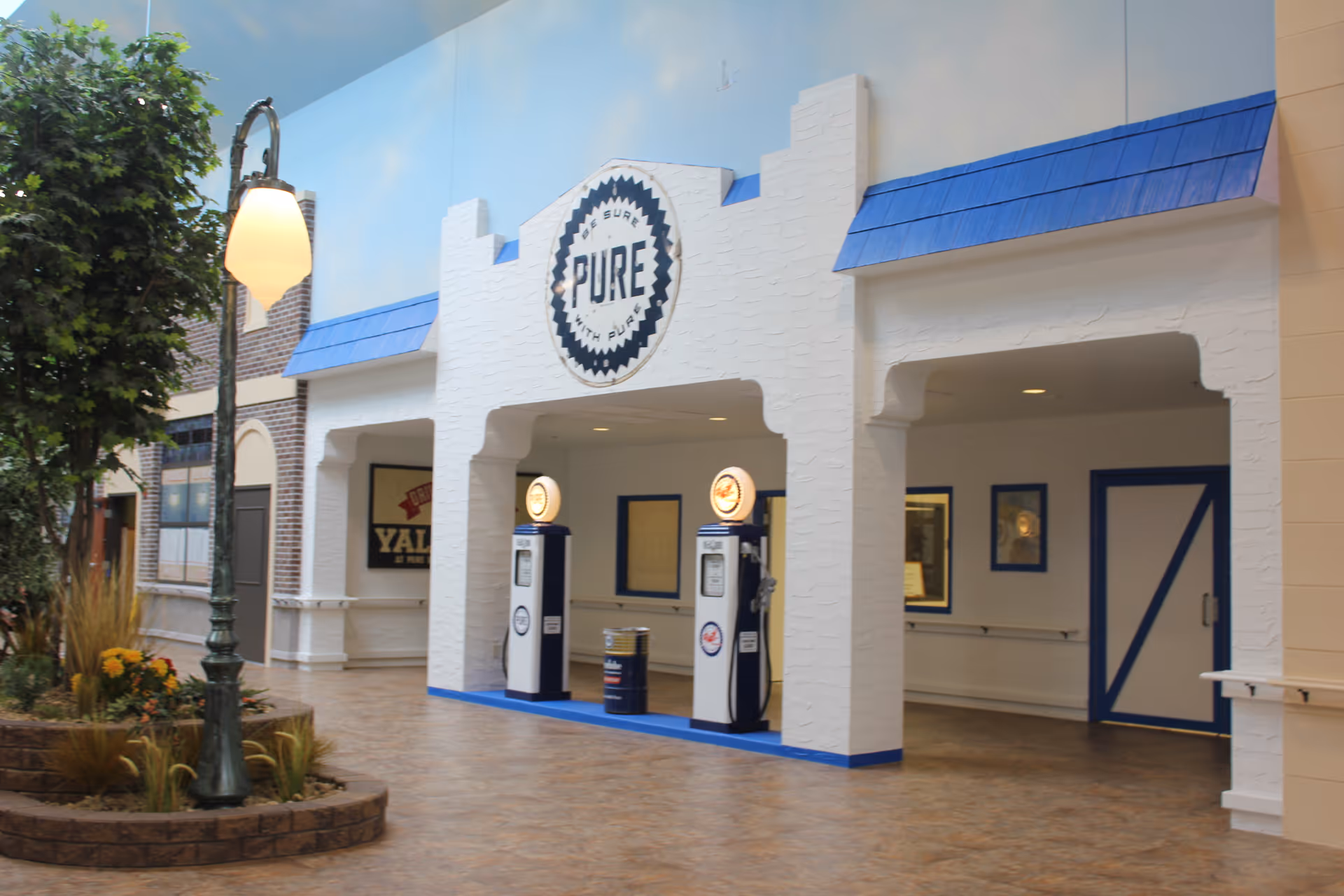 Indoor area designed to look like an old-fashioned street scene with a white building facade featuring blue trim and two vintage-style gas pumps. There is a street lamp and a small planted area with greenery and flowers on the left side.