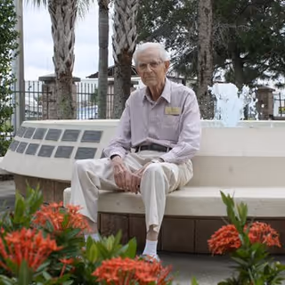 An elderly man wearing a light purple striped shirt and beige pants sits on a curved white bench in an outdoor garden area with red flowers in the foreground, palm trees, and a water fountain in the background.