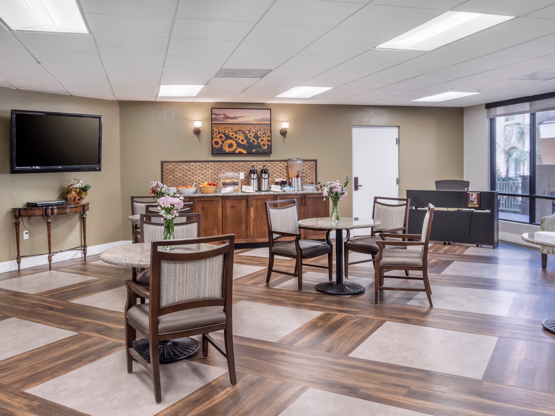 Bright common dining area with round tables and chairs, a buffet counter, wall TV and floral centerpieces.