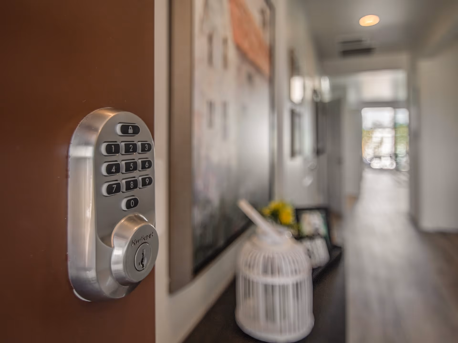 Close-up of a silver keypad door lock on a brown door with a blurred hallway and decorative items in the background.