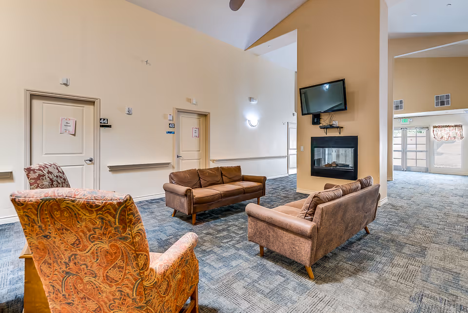 A senior living facility common area with two brown sofas and an orange patterned armchair arranged around a wall-mounted TV and fireplace. The room has beige walls, blue patterned carpet, and doors labeled 44 and 45. There is a large mirror on one wall and an exit door with a window and floral valance in the background.