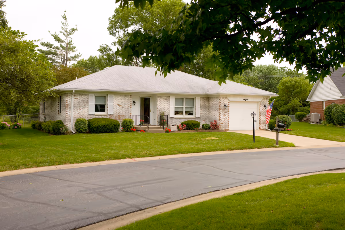 Front view of a single-story brick house with a garage, driveway, and well-kept lawn under leafy trees.