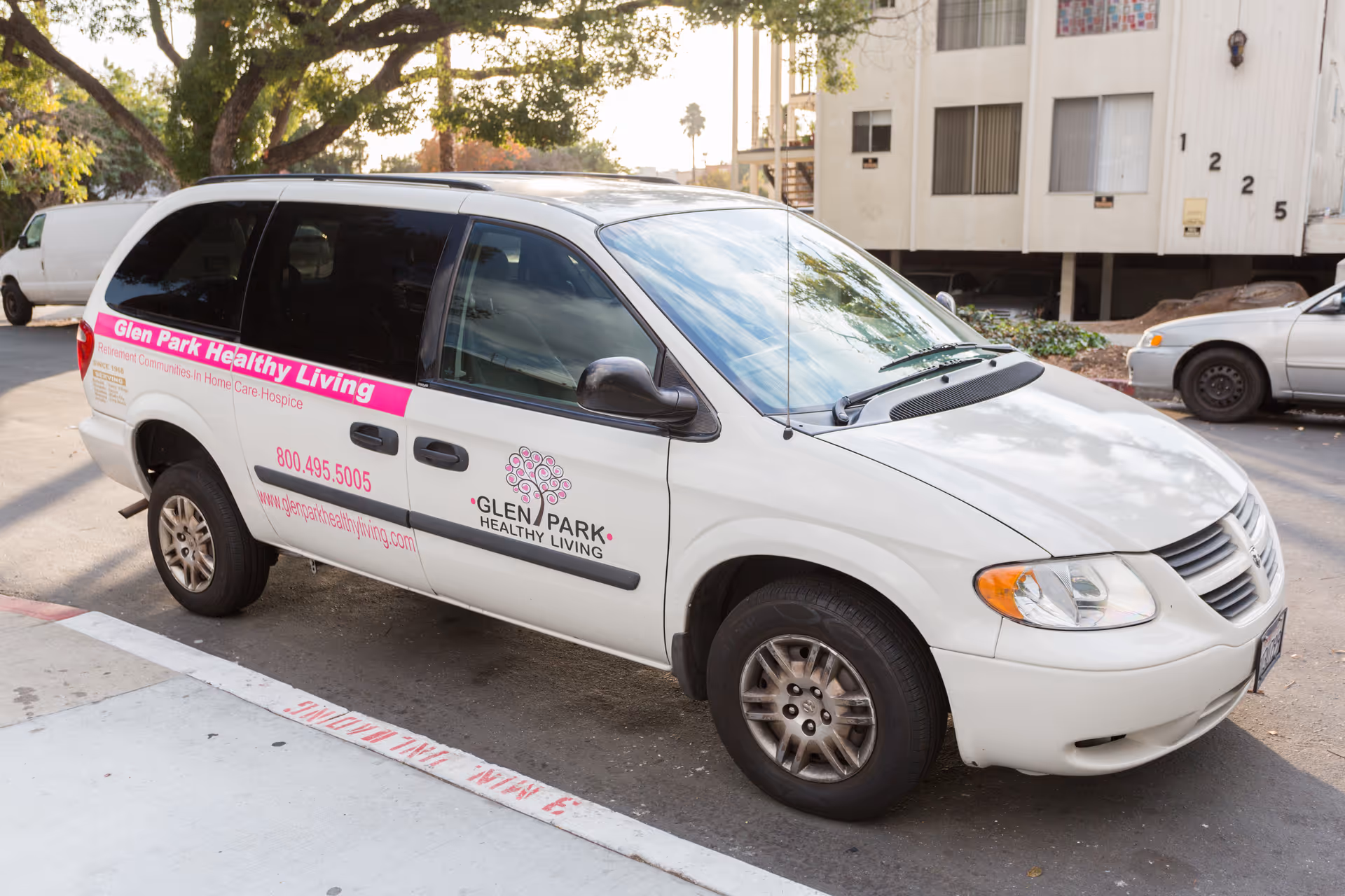 White 'Glen Park Healthy Living' minivan parked on a street in front of an apartment building.