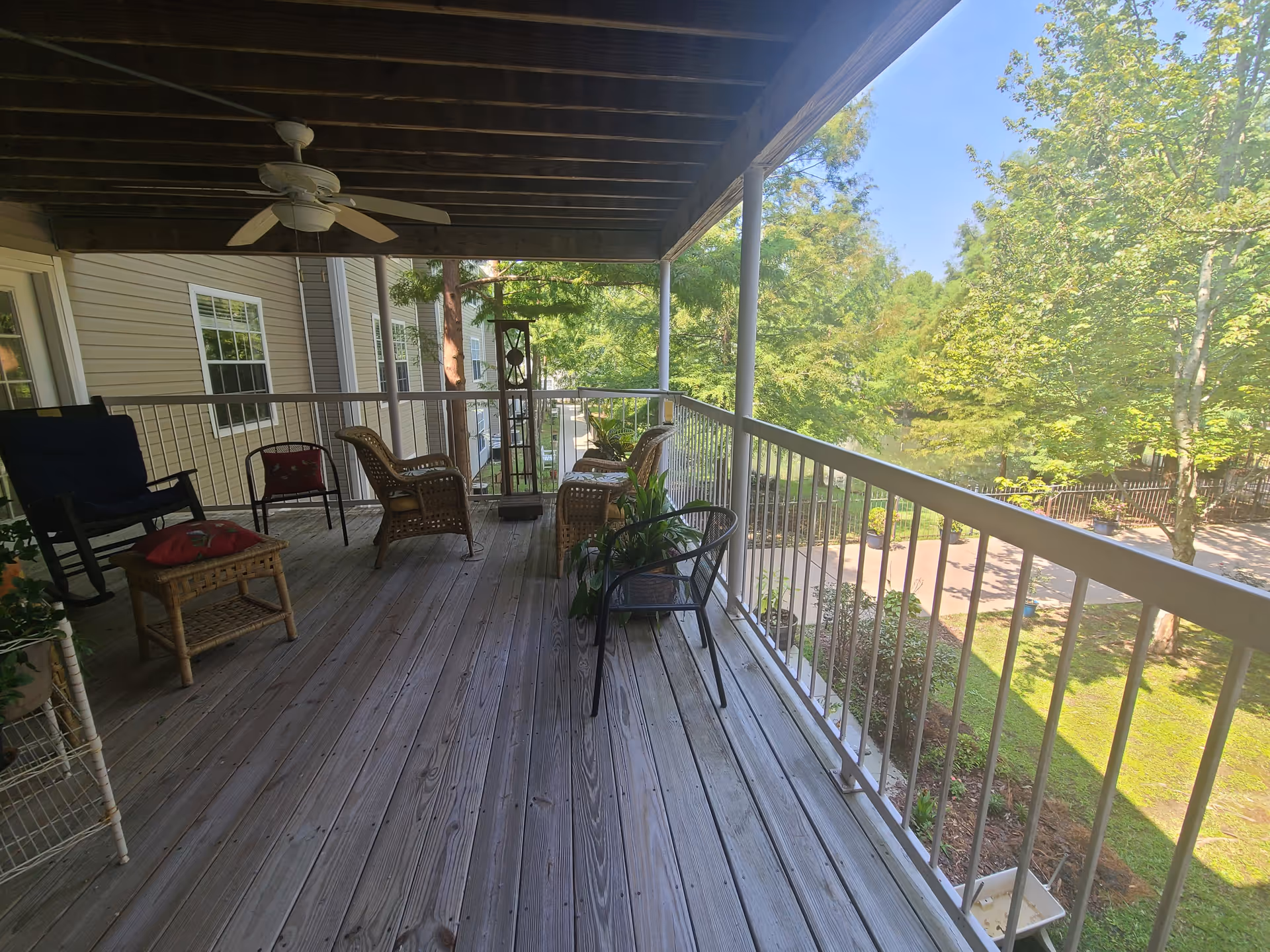 Covered wooden balcony with various chairs including wicker and metal, a ceiling fan, potted plants, and a view of trees and a garden area outside.