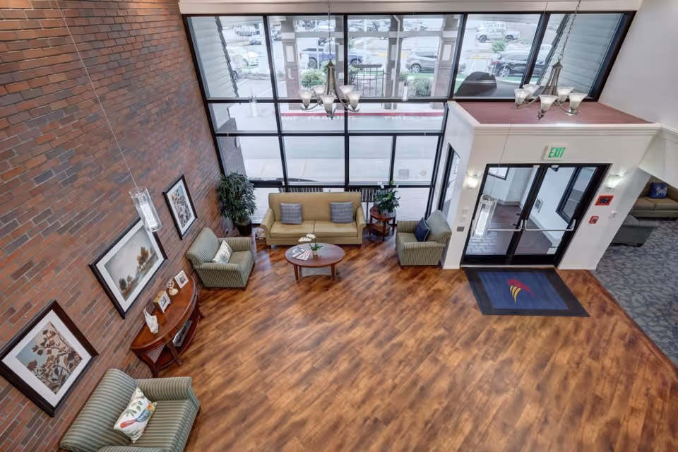 A spacious senior living facility lobby with wooden flooring, a brick accent wall, and large floor-to-ceiling windows. The seating area includes a beige sofa, two striped armchairs, a round wooden coffee table, and side tables with plants. There are framed artworks on the brick wall and a chandelier hanging from the ceiling. The entrance door has an exit sign above it and a blue mat with a colorful logo on the floor.