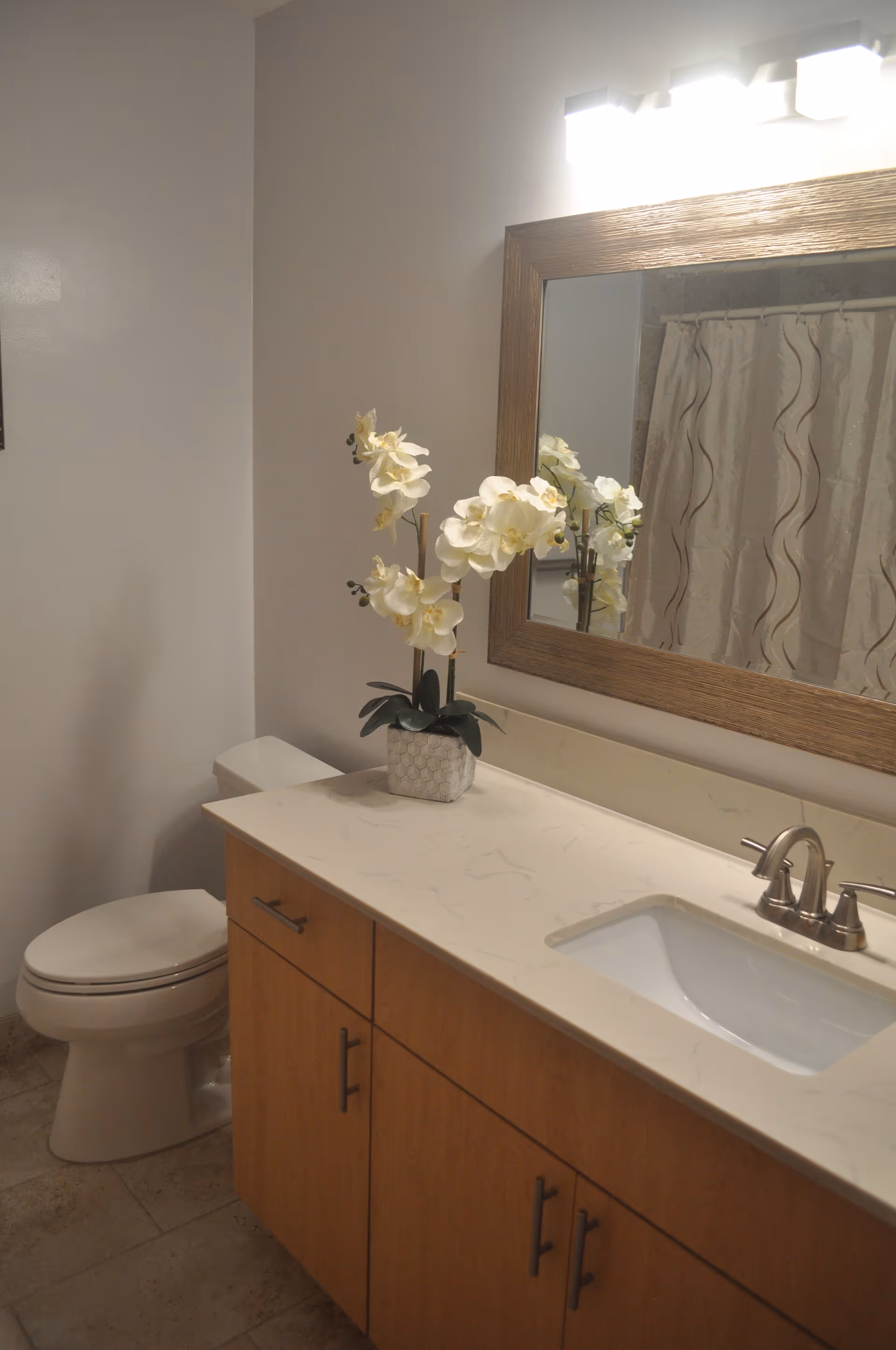 A bathroom with a white toilet, a wooden vanity with a white countertop and built-in sink, a silver faucet, a large rectangular mirror with a wooden frame, and a decorative white orchid plant on the countertop. A shower curtain with a wavy pattern is visible in the reflection of the mirror.