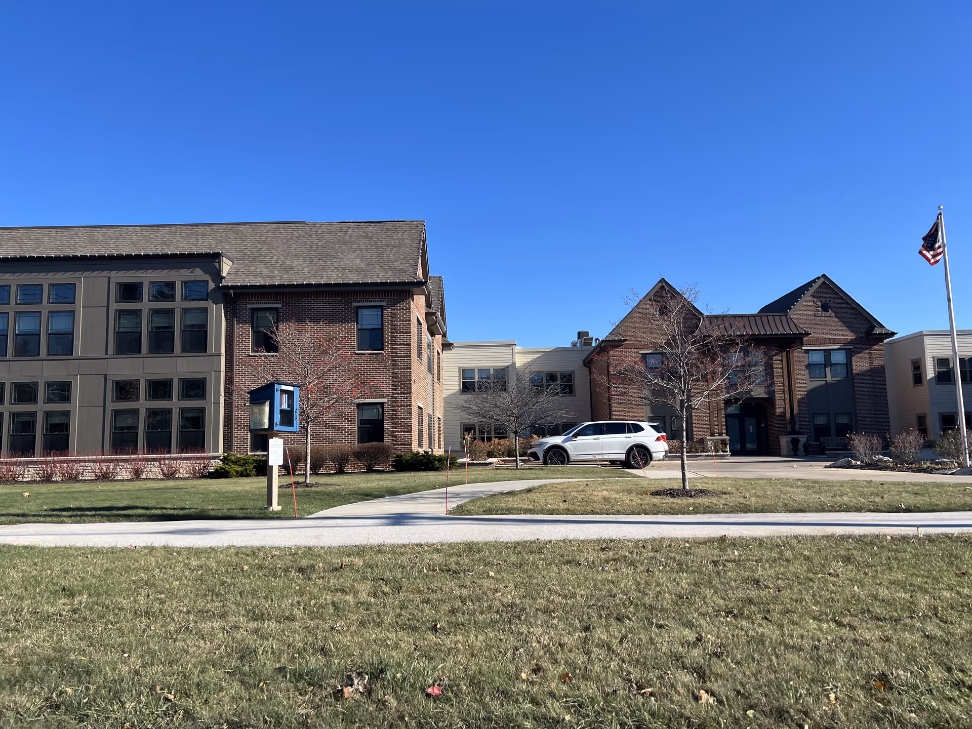 Exterior view of Azura Assisted Living and Memory Care of Fox Point building on a clear day, showing a brick and siding facade with multiple windows, a white SUV parked in front, a flagpole with an American flag, and a grassy lawn with a sidewalk leading to the entrance.