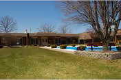 Outdoor view of a senior living facility with a large grassy lawn, a swimming pool surrounded by a stone wall, and a single-story brick building in the background under a clear blue sky.