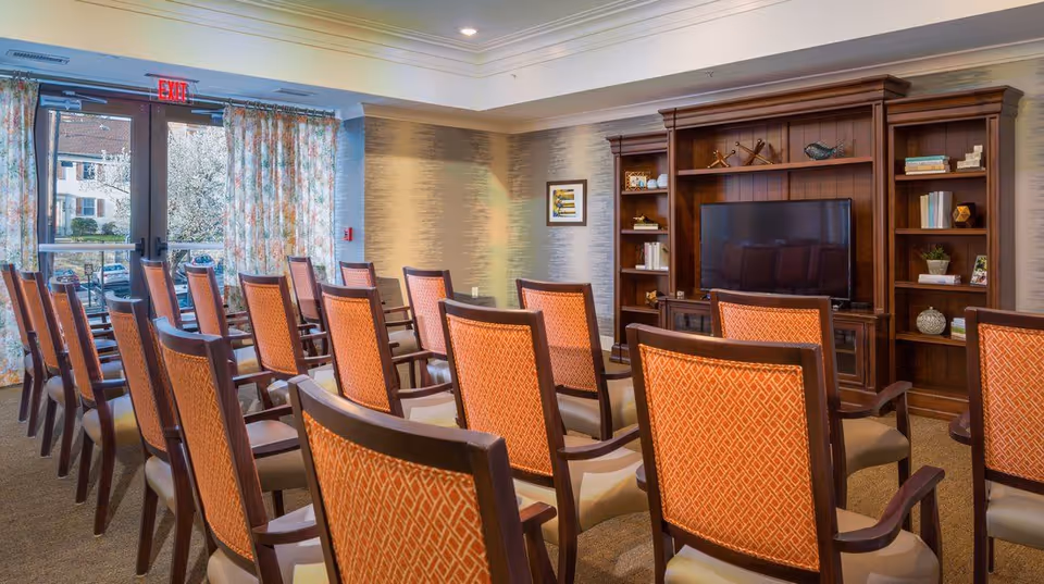 A well-lit room with multiple rows of wooden chairs featuring orange patterned upholstery, facing a large wooden entertainment center with a flat-screen TV and decorative items on shelves. The room has floral curtains covering glass doors that lead outside, and a framed picture on the wall.